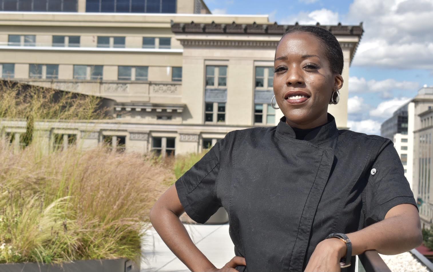 Jazmine Moore, founder of The Invisible Host, wearing a black chef coat in an outdoor city setting.